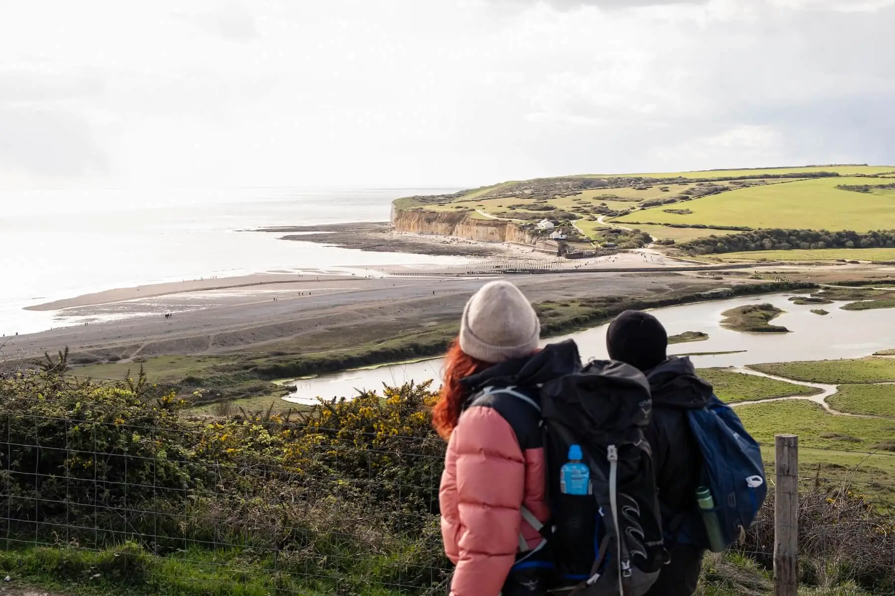 Two ramblers overlook the South Downs with cliffs, fields, and a winding river below.