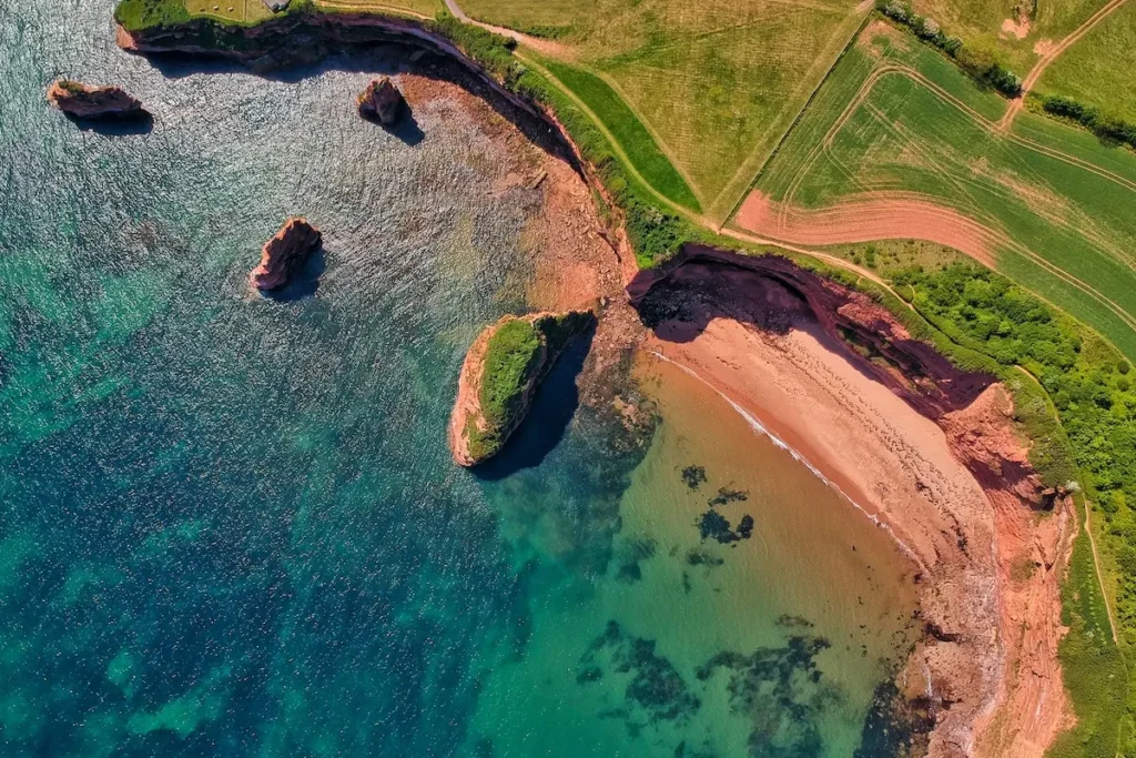Aerial view of rocky coastline, clear blue water, sandy beach, green fields, and circular walks.